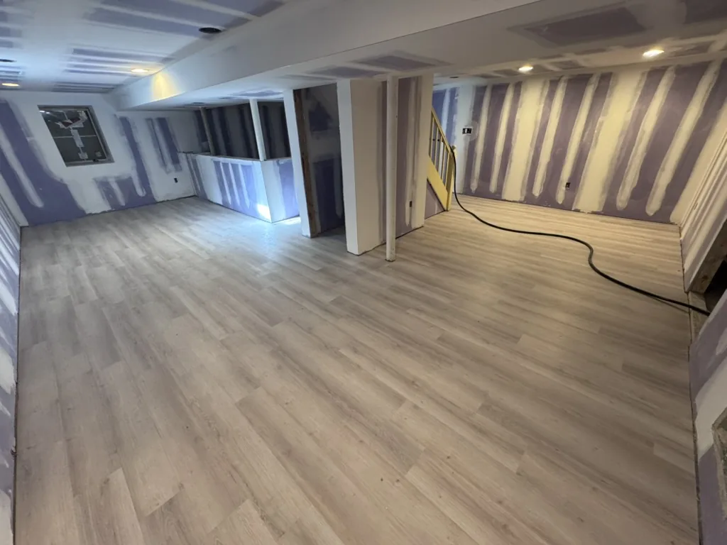 LVT plank flooring installed across a basement room, with taped drywall, recessed lighting and a stair rail at the back