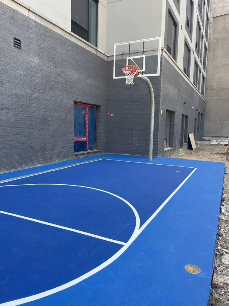 Blue basketball half court between two apartment buildings, with a glass backboard on a pole against a dark brick wall