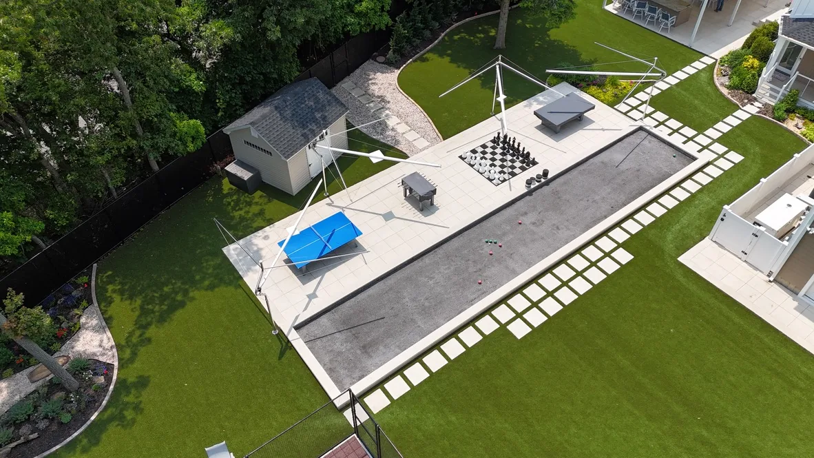 Aerial view of the outdoor gaming area with the sail shades furled, showing the bocce court, giant chess board, shuffleboard and ping pong table on a paver patio surrounded by turf