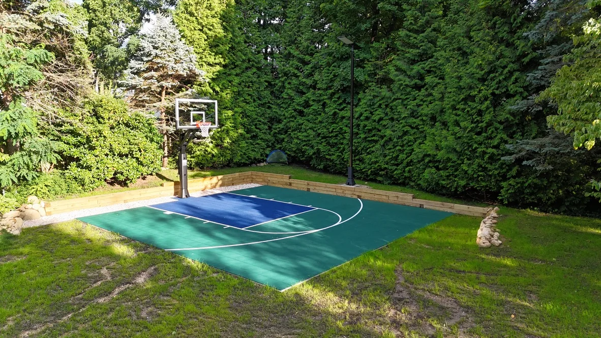 Green and blue backyard half court with a hoop, timber retaining wall, and light pole