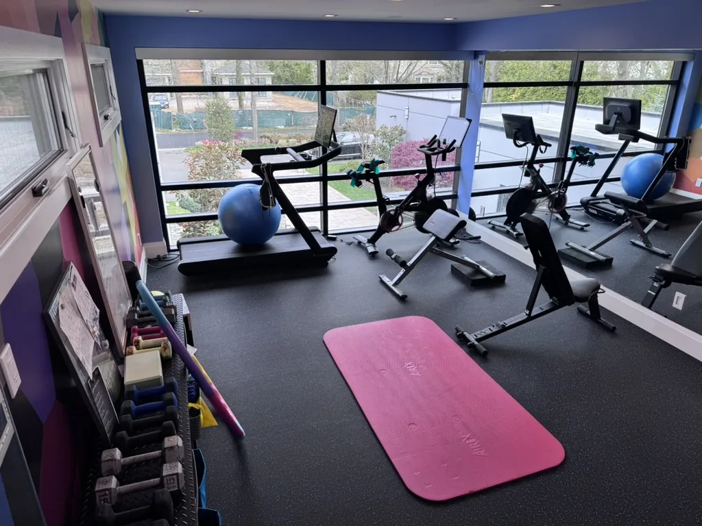 Rubber gym flooring in a home fitness room with a treadmill, spin bikes, dumbbells and exercise balls beside a wall of windows