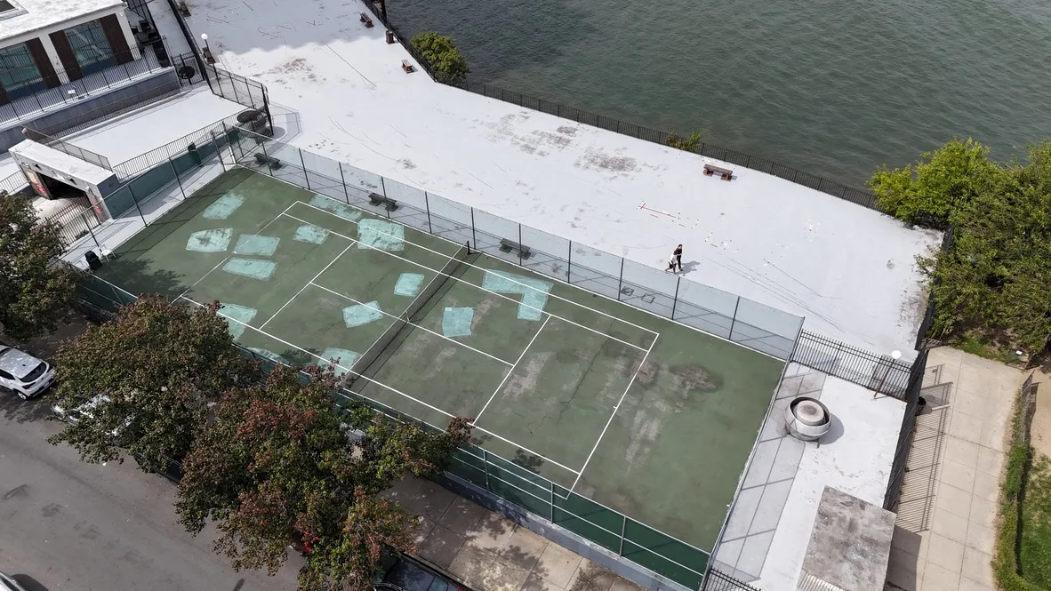 The community tennis court before the work, with a faded and patched green surface inside chain link fencing beside the water
