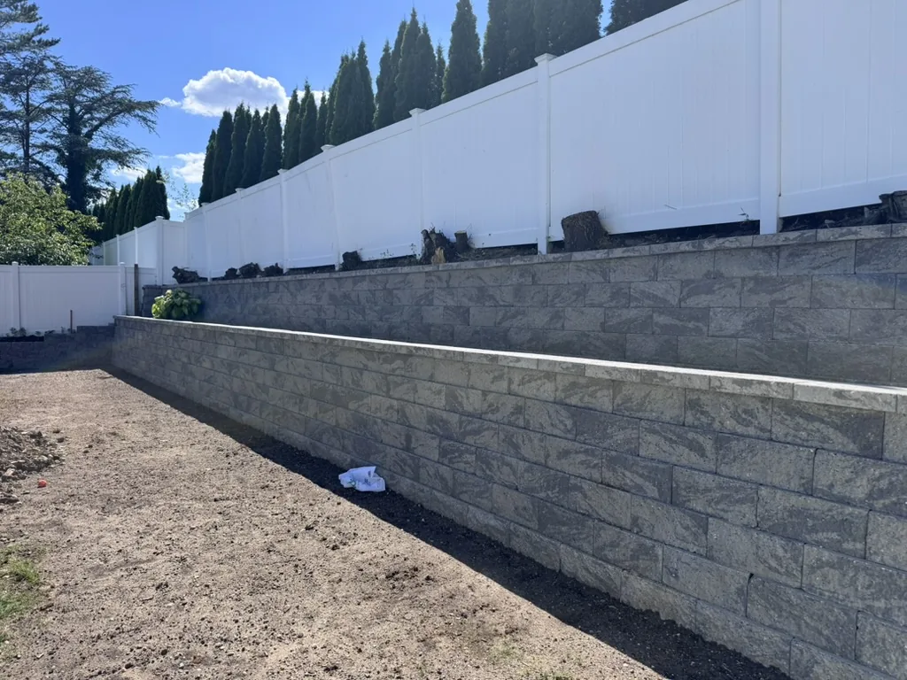 The rebuilt wall in two tiers of grey segmental block with a capped top course, below the same white vinyl fence