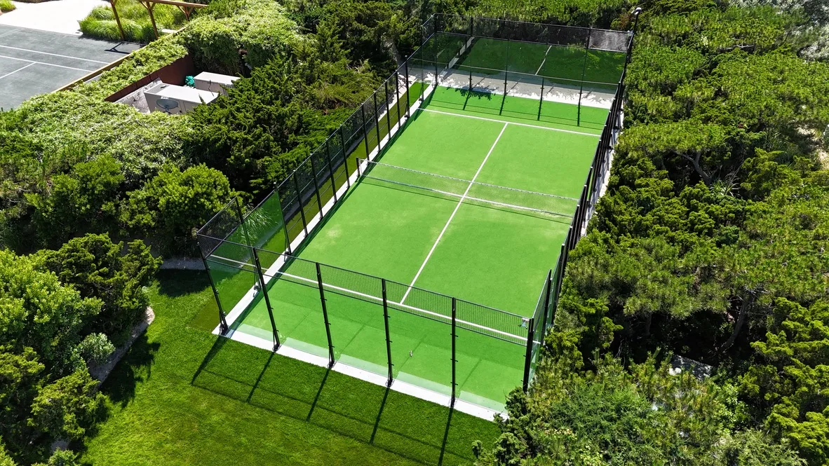 Aerial view of a padel court with glass and mesh walls and a green turf surface, set into landscaped grounds beside a tennis court