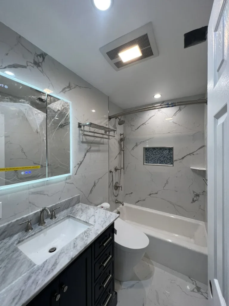 Renovated bathroom with marble effect porcelain tile, a lit mirror, a dark vanity with a marble top and a tub shower with a mosaic niche