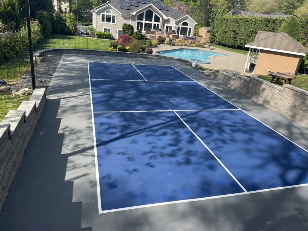 Blue pickleball court with white lines on a grey surround, set below a pool and house on a terraced lawn