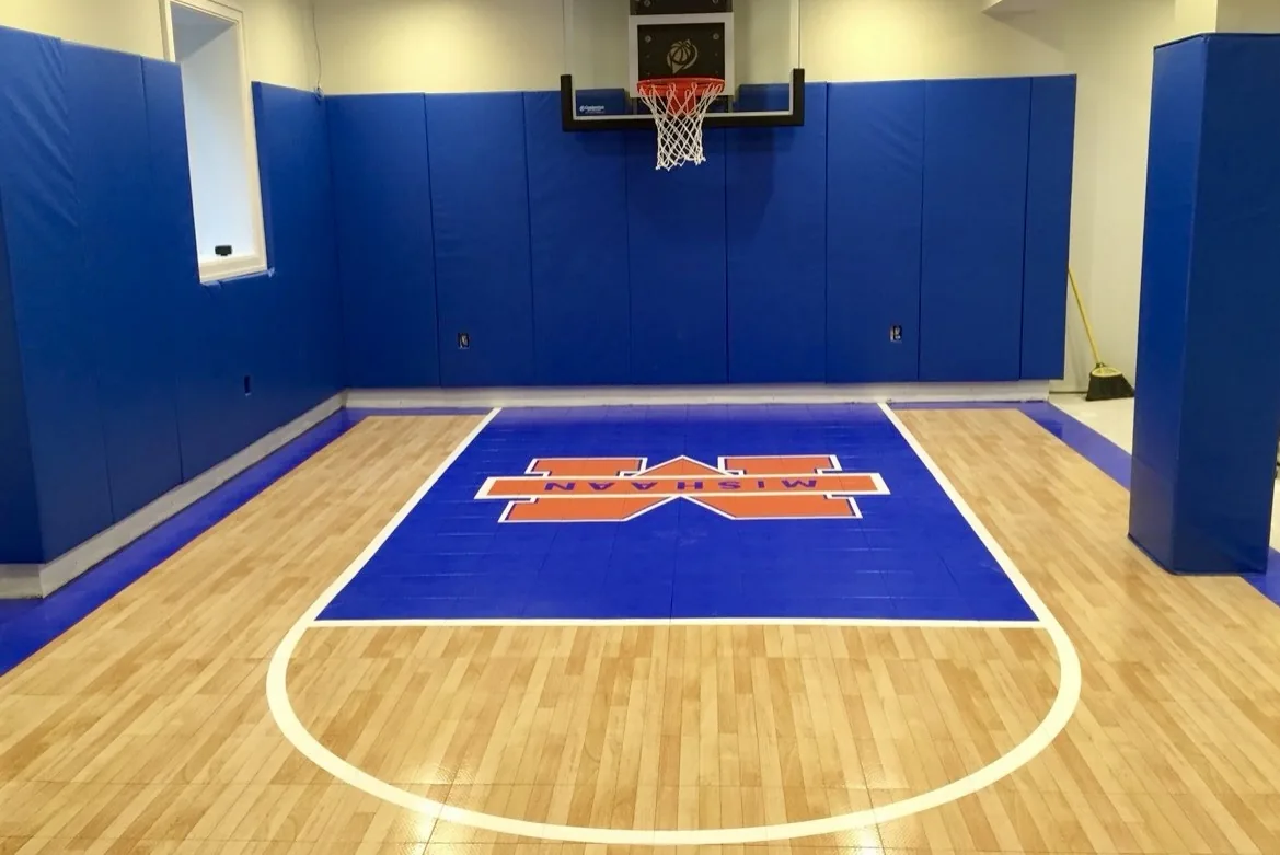 Indoor basketball court with blue padded walls, a wall mounted hoop and a wood look tile surface with a custom painted centre logo