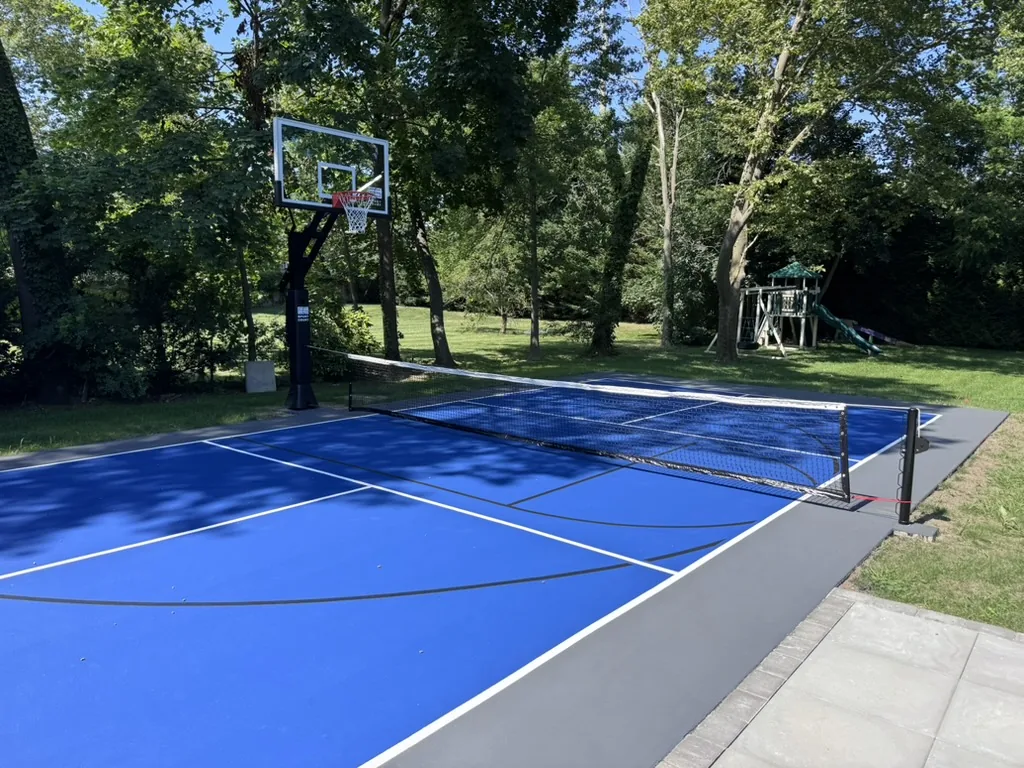 Blue multisport court with a basketball hoop and a pickleball net, set in a wooded backyard beside a play set
